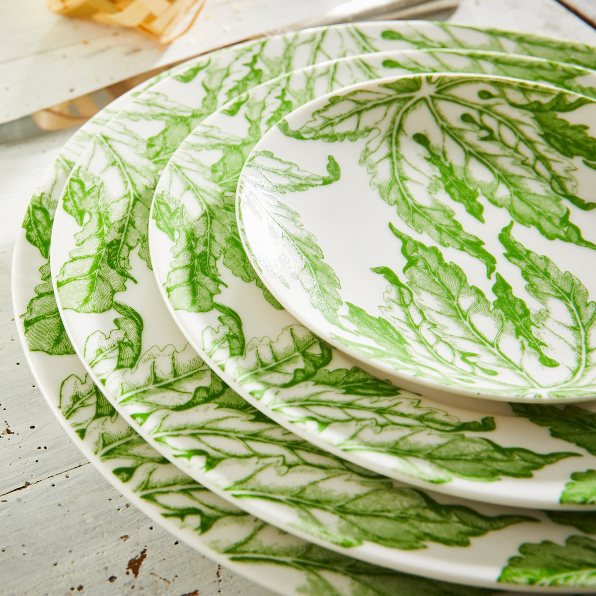 A set of white porcelain Freya Rimmed Charger Plates by Caskata Artisanal Home, adorned with green leaf patterns, arranged in a stack on a rustic white wooden surface.