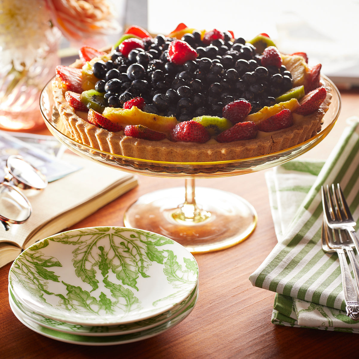 Fruit tart topped with blueberries, strawberries, and kiwi on a glass stand. Surrounding items include heirloom-quality dinnerware, Caskata Artisanal Home&#39;s Freya Small Plates with a green floral pattern, fork and knife, glasses, book, and a green striped napkin.
