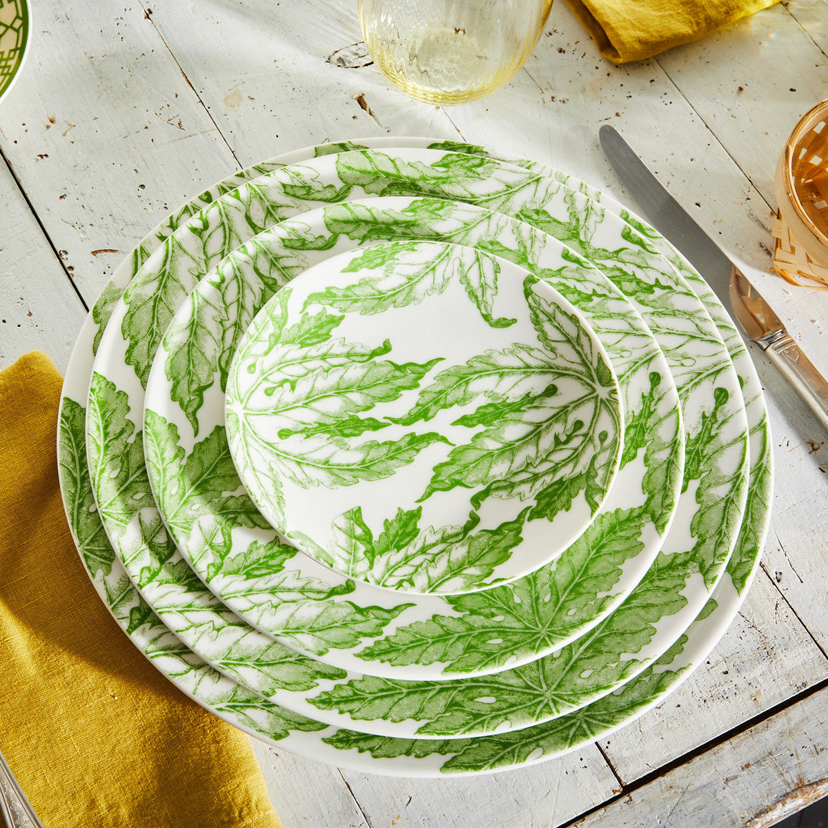 A set of Caskata Artisanal Home Freya Rimmed Charger Plates with green leaf patterns stacked on a rustic wooden table, accompanied by a yellow napkin, a knife, and glassware, exuding the charm of green florals.