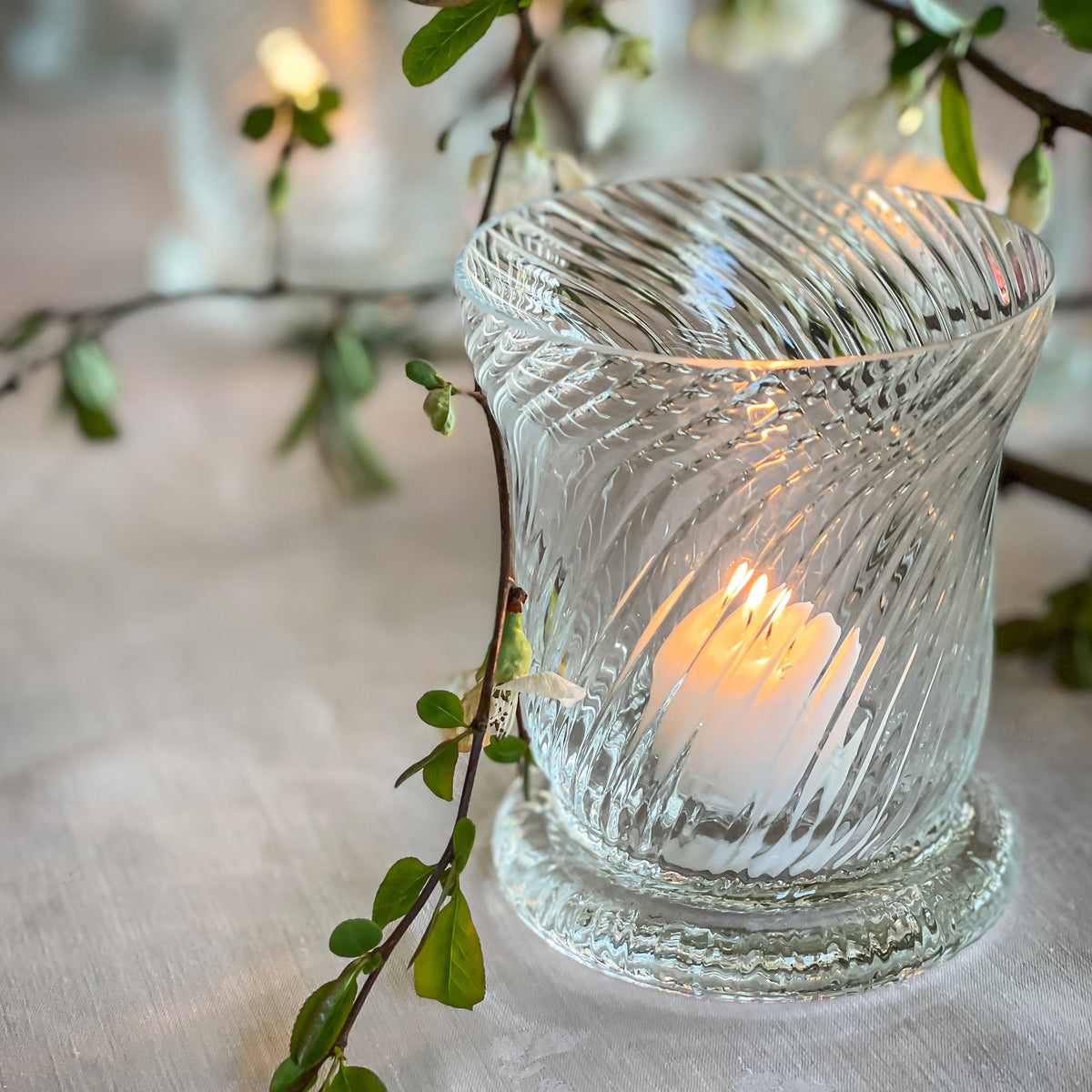 A Quinn Clear Votive by Caskata holds a lit candle and is surrounded by leafy green branches on a spring tabletop.