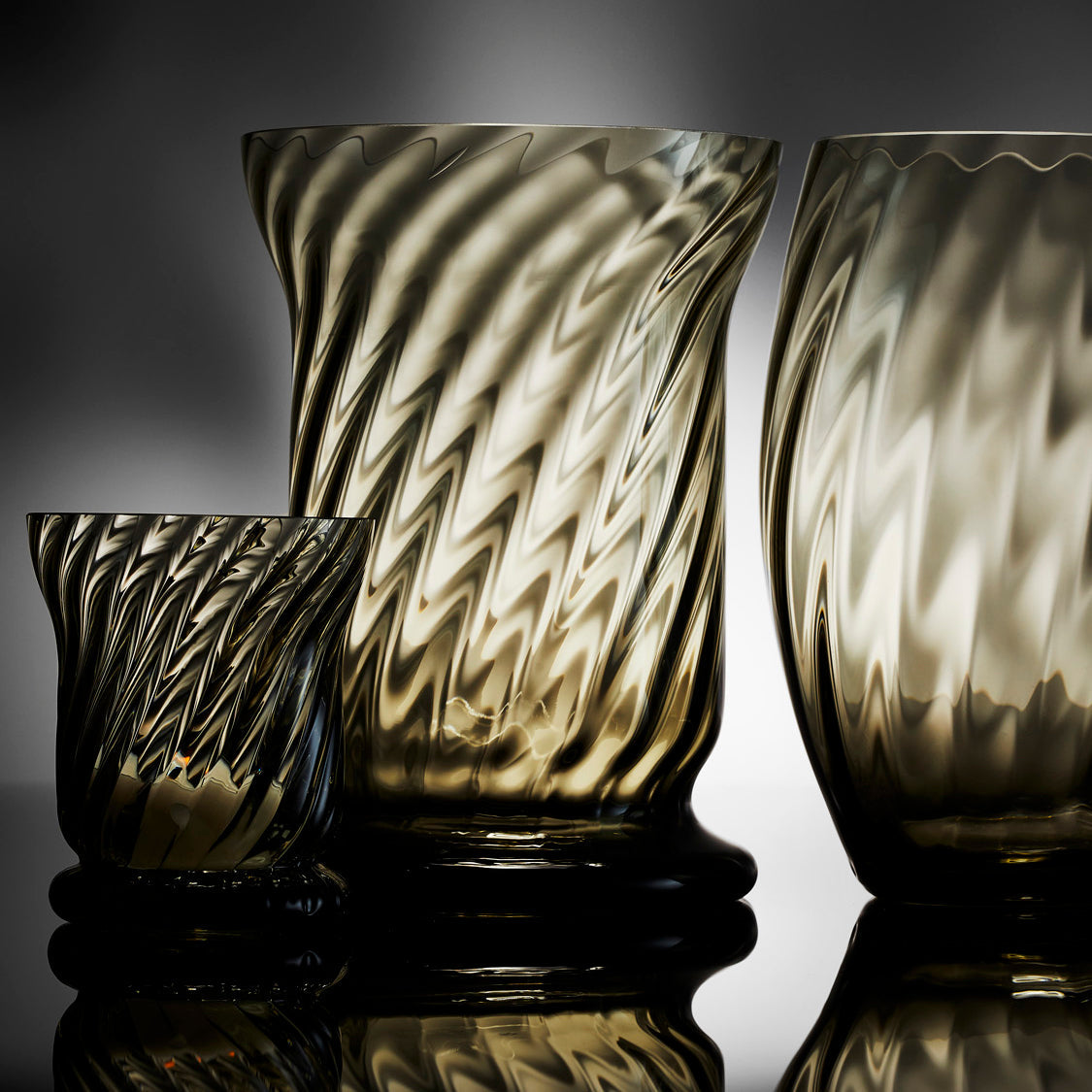 The Quinn Smoke Optics Collection features a Votive and Boothbay and Elliot Hurricanes. Swirling glass optics and smokey glass make these candle holders from Caskata perfect for every table.