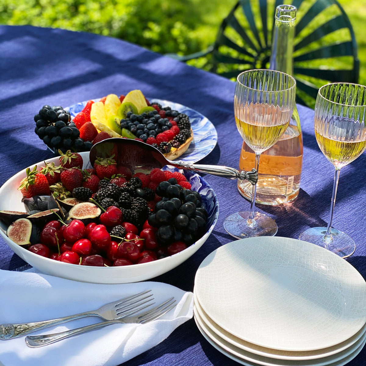 A summer picnic setup featuring a bowl of mixed berries and figs in a Caskata Artisanal Home Peony Wide Serving Bowl, with two glasses of rosé wine, on a table with a blue-striped tablecloth.