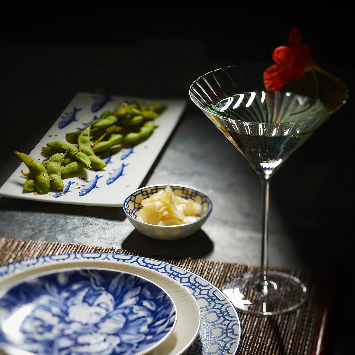 A cocktail garnished with a red flower is on a table beside a plate of edamame, a small bowl of pickled ginger, and a patterned dish featuring an interlocking lattice pattern from the Newport Dipping Dishes, Set of 4 by Caskata.