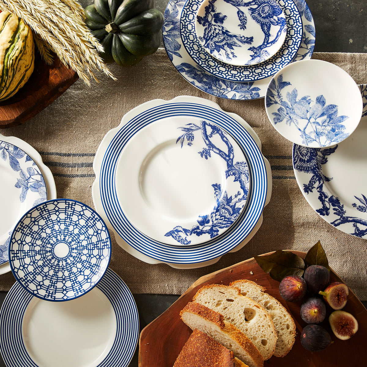 A variety of heirloom-quality dinnerware with blue and white patterns is arranged on a beige tablecloth, next to bread, figs, and a couple of gourds, including Arbor Blue Small Plates by Caskata Artisanal Home.
