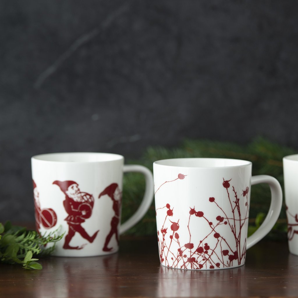 Three **Elves Mugs**, part of a whimsical holiday collection by **Caskata Artisanal Home**, are decorated with red designs of playful elves carrying gifts and botanical patterns. They rest on a dark wooden surface with green foliage in the background.