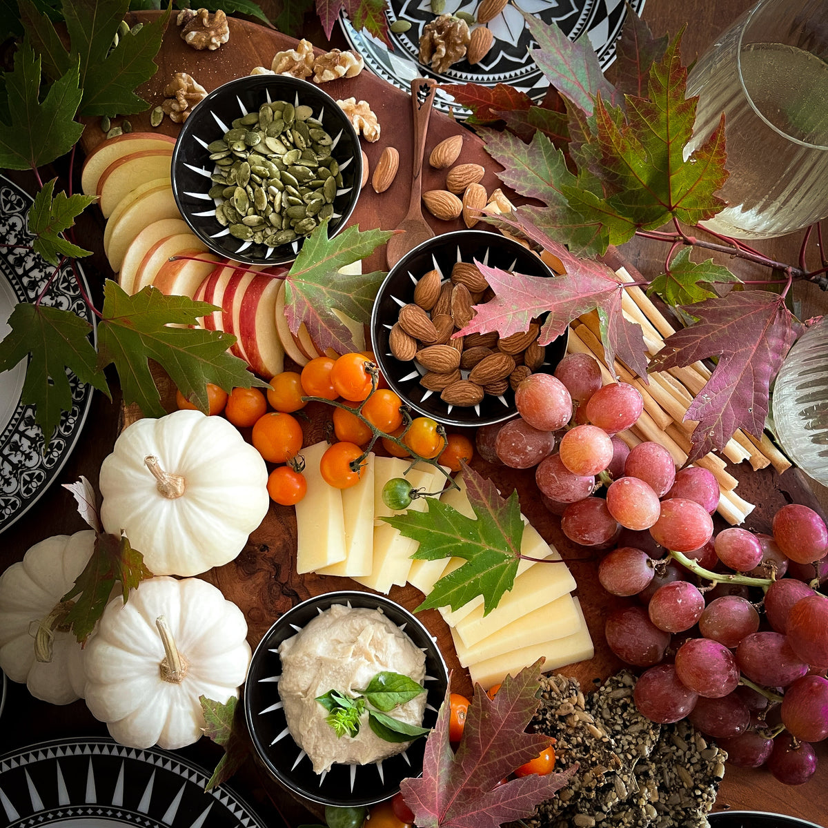 A wooden board with grapes, cheese, nuts, seeds, and apple slices is beautifully arranged among colorful autumn leaves. Tiny white pumpkins and Caskata Marrakech Dipping Dishes, Set of 4 complete the scene.