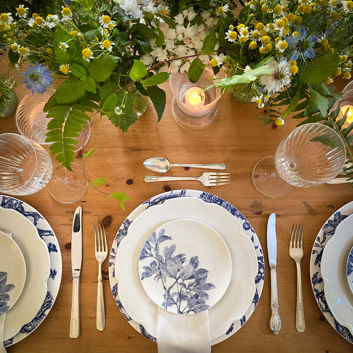 A wooden table set for a meal with Caskata Artisanal Home Arbor Blue Small Plates, heirloom-quality dinnerware, silver cutlery, crystal glasses, lighted candles, and a centerpiece of assorted flowers and greenery.