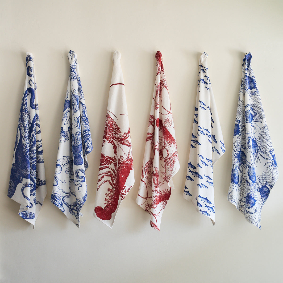 Six Lucy Kitchen Towels, Set of 2 by Caskata in blue and red hues hang neatly on hooks against a white wall.