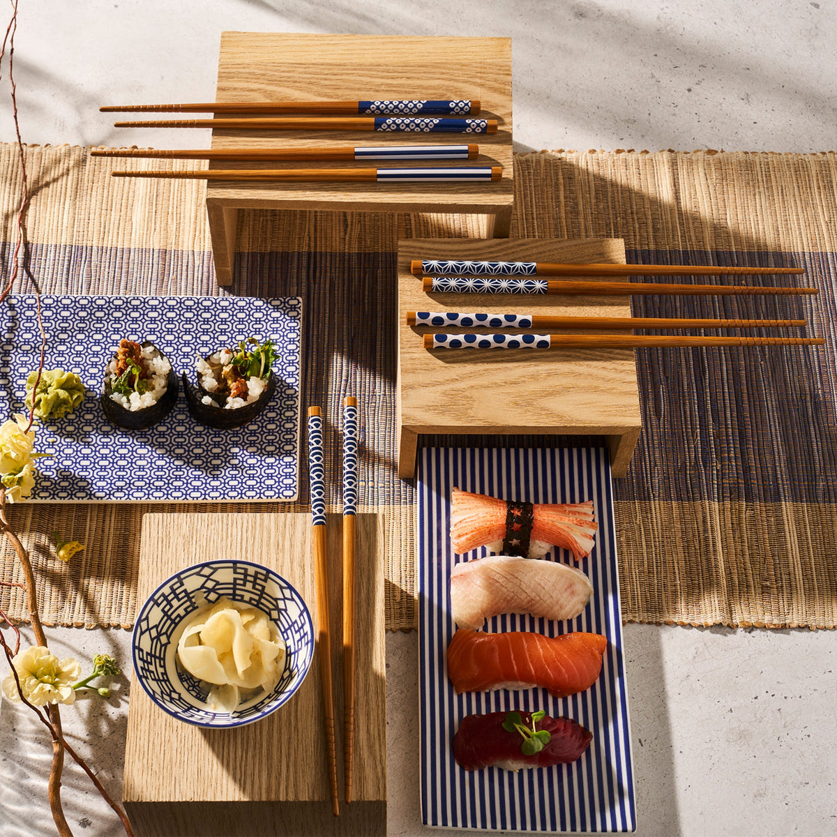 A table set with sushi, nigiri, and small bowls of pickled ginger on patterned plates, including a premium Caskata Newport Large Sushi Tray. Wooden chopsticks rest on holders alongside the dishes. The setup is completed with a bamboo mat and wooden platform.