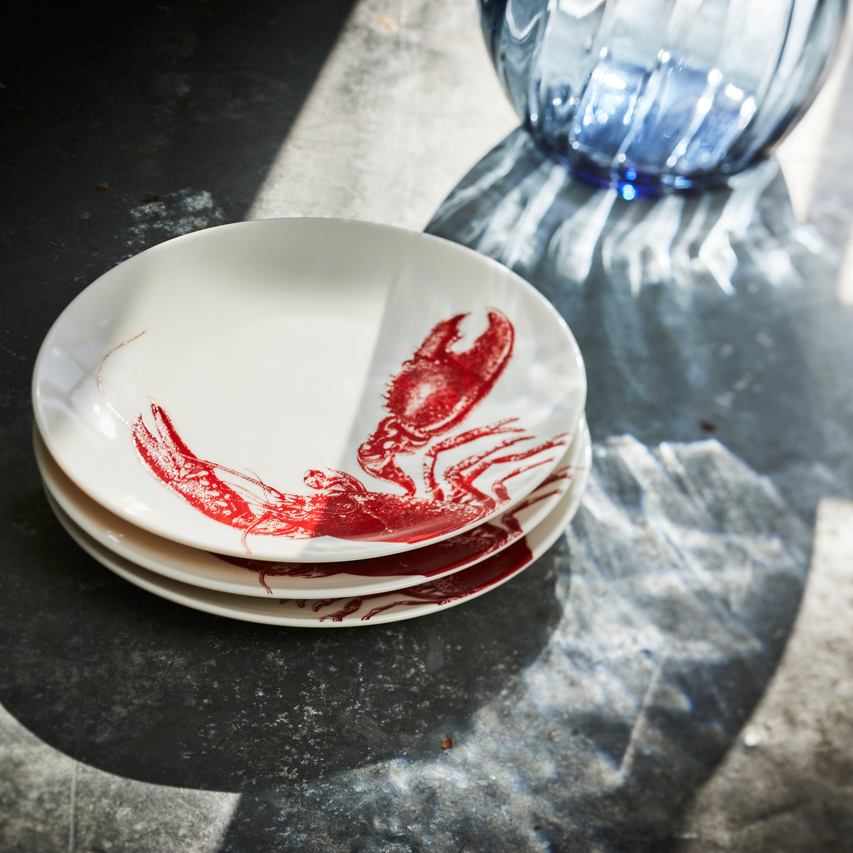 Stack of Caskata Artisanal Home Lobster Red Small Plates featuring red lobster illustrations, placed on a reflective surface with a blue glass vase nearby. Sunlight casts shadows across the scene, adding to the seaside style ambiance.