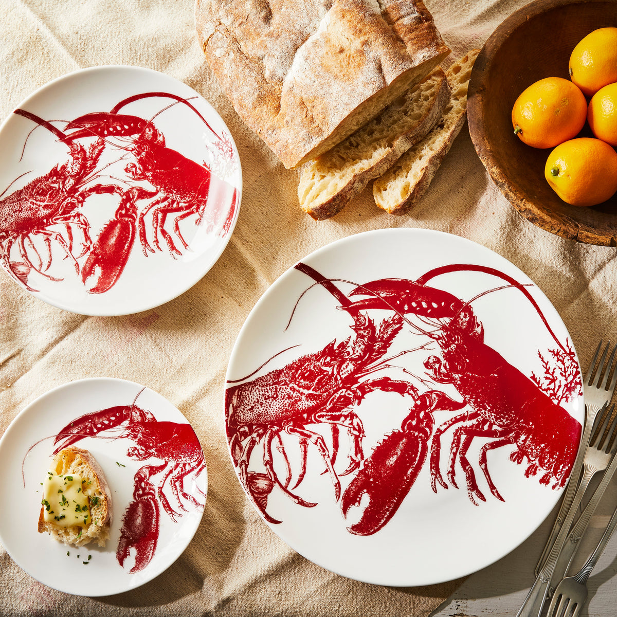 Three Lobster Coupe Dinner Plates by Caskata, crafted from premium porcelain and dishwasher and microwave safe, a loaf of bread, a wooden bowl of lemons, and silverware are arranged on a beige tablecloth.