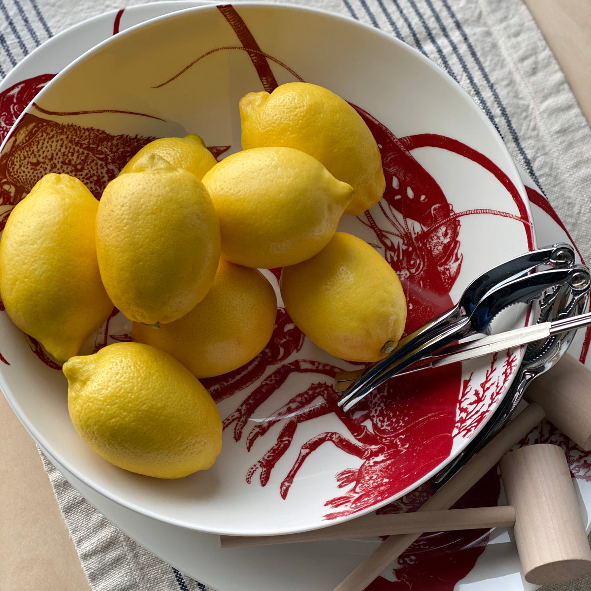 A white Lobster Wide Serving Bowl from Caskata Artisanal Home with a red lobster illustration holds several yellow lemons, a lemon squeezer, and a fork on a striped cloth background, capturing the essence of seaside style tableware.