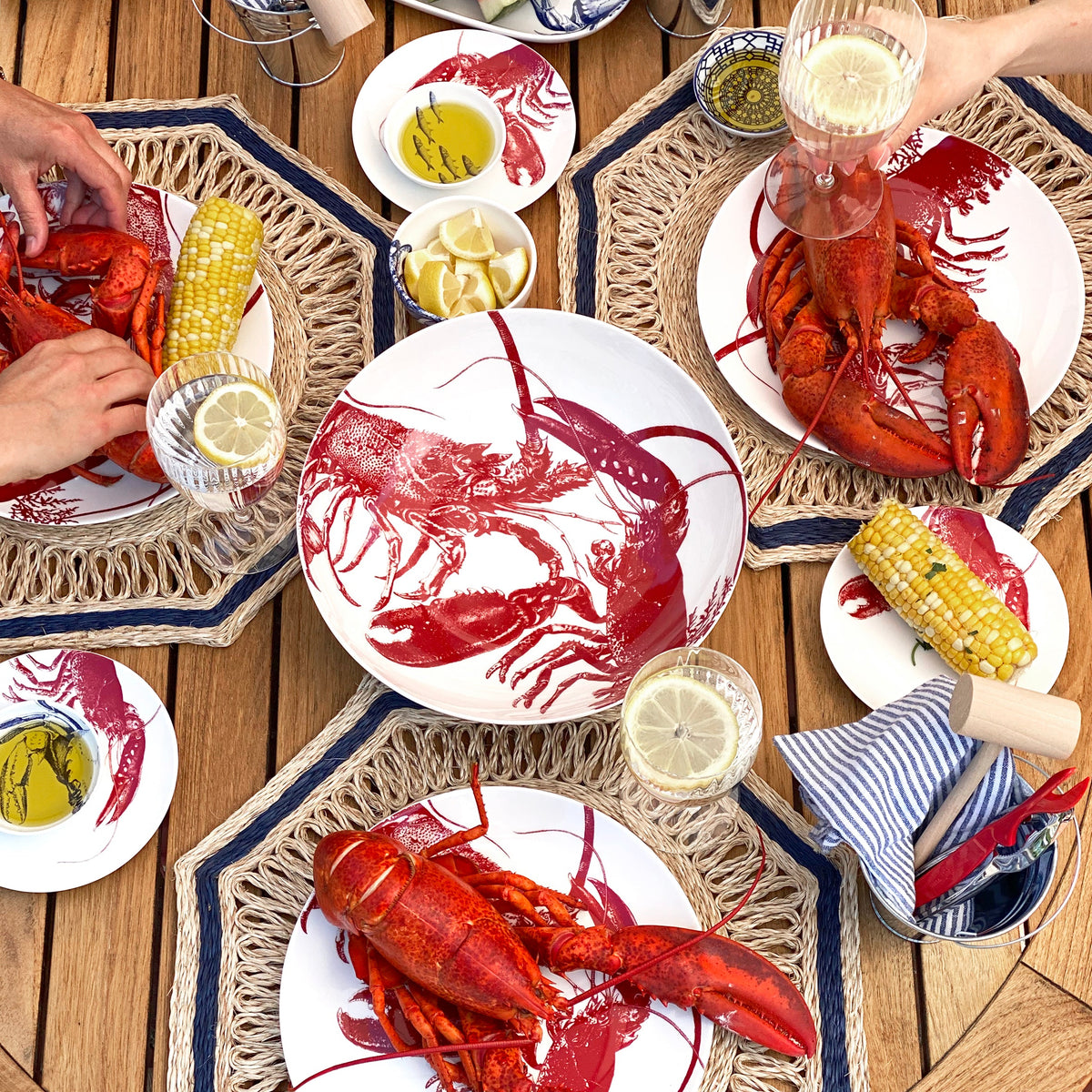 Four premium Caskata Lobster Coupe Dinner Plates paired with corn on the cob and lemon wedges on a wooden table. Three hands can be seen, one holding a glass of lemonade. The scene is set on nautical-themed placemats, showcasing these dishwasher and microwave safe seaside style dinner plates.