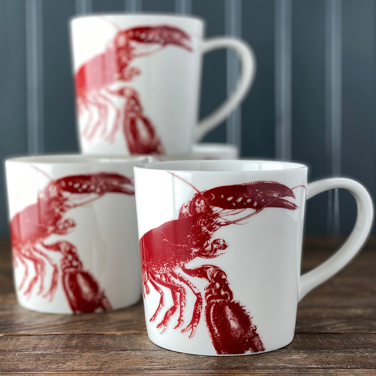 Three Caskata Artisanal Home Lobster Red Mugs featuring red lobster illustrations are stacked on a wooden surface against a vertically striped dark background, embodying a charming seaside style.