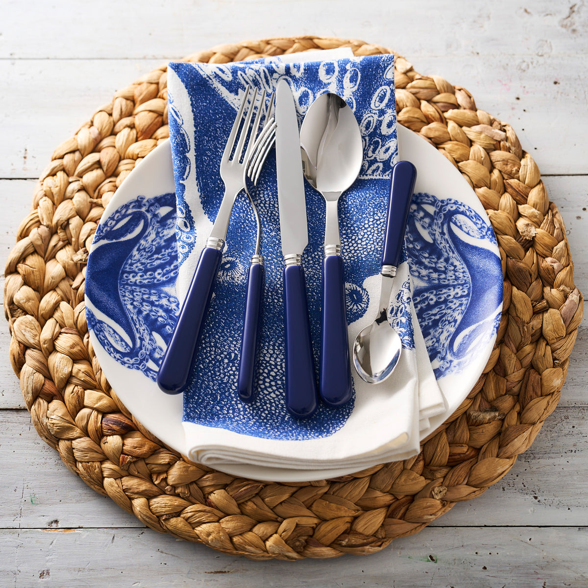 A place setting with a creamy white Lucy Coupe Dinner Plate from Caskata Artisanal Home, blue-handled cutlery, a blue-patterned napkin, and a braided placemat on a white wooden table.