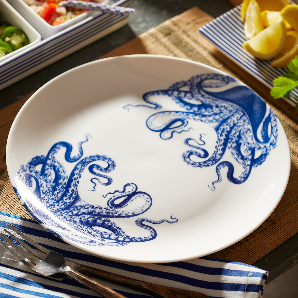 A creamy white premium porcelain Lucy Coupe Dinner Plate by Caskata Artisanal Home features blue octopus tentacles on two sides, placed on a brown placemat alongside a silver fork, knife, and a blue striped napkin. Background includes lemon slices and a portion of food in a dish—meet Lucy the octopus!