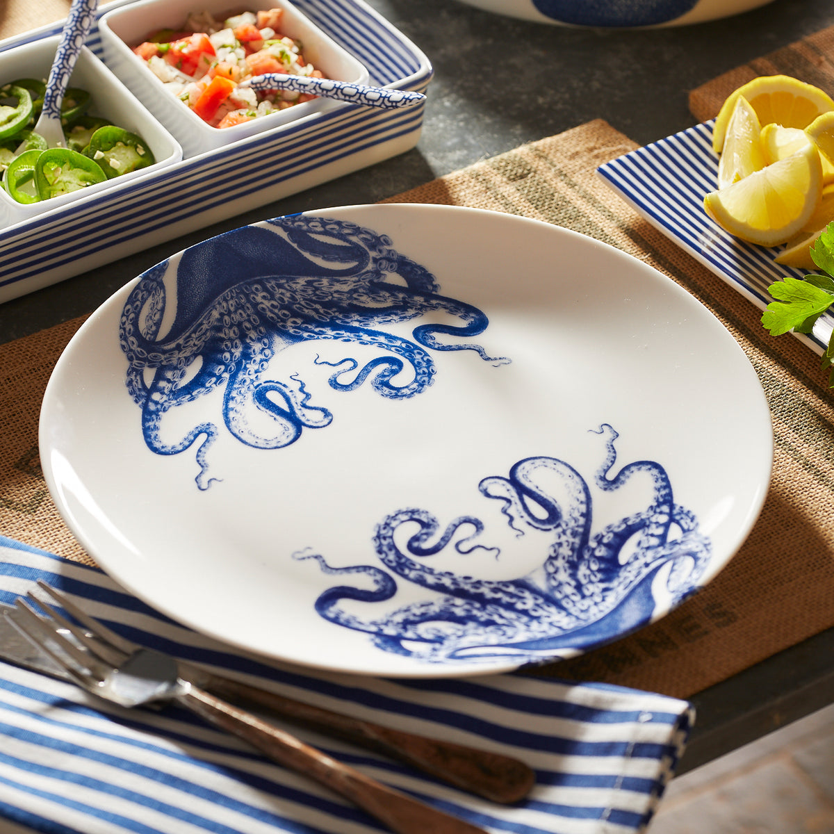 A creamy white premium porcelain Lucy Coupe Dinner Plate by Caskata Artisanal Home with a blue Lucy the octopus design is set on a table with a blue and white striped napkin, fork, knife, and bowls of lemon, sliced jalapenos, and diced vegetables.