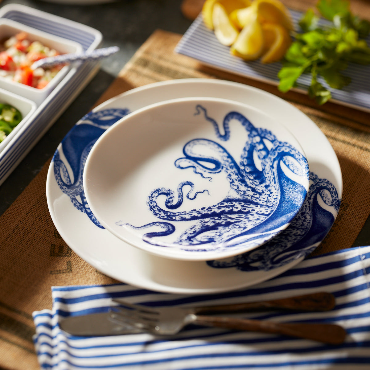 A creamy white premium porcelain Lucy Coupe Dinner Plate from Caskata Artisanal Home with a blue octopus design, named Lucy the octopus, rests on a matching bowl. The setting features a blue striped napkin, silverware, and a dish of lemons and herbs in the background.