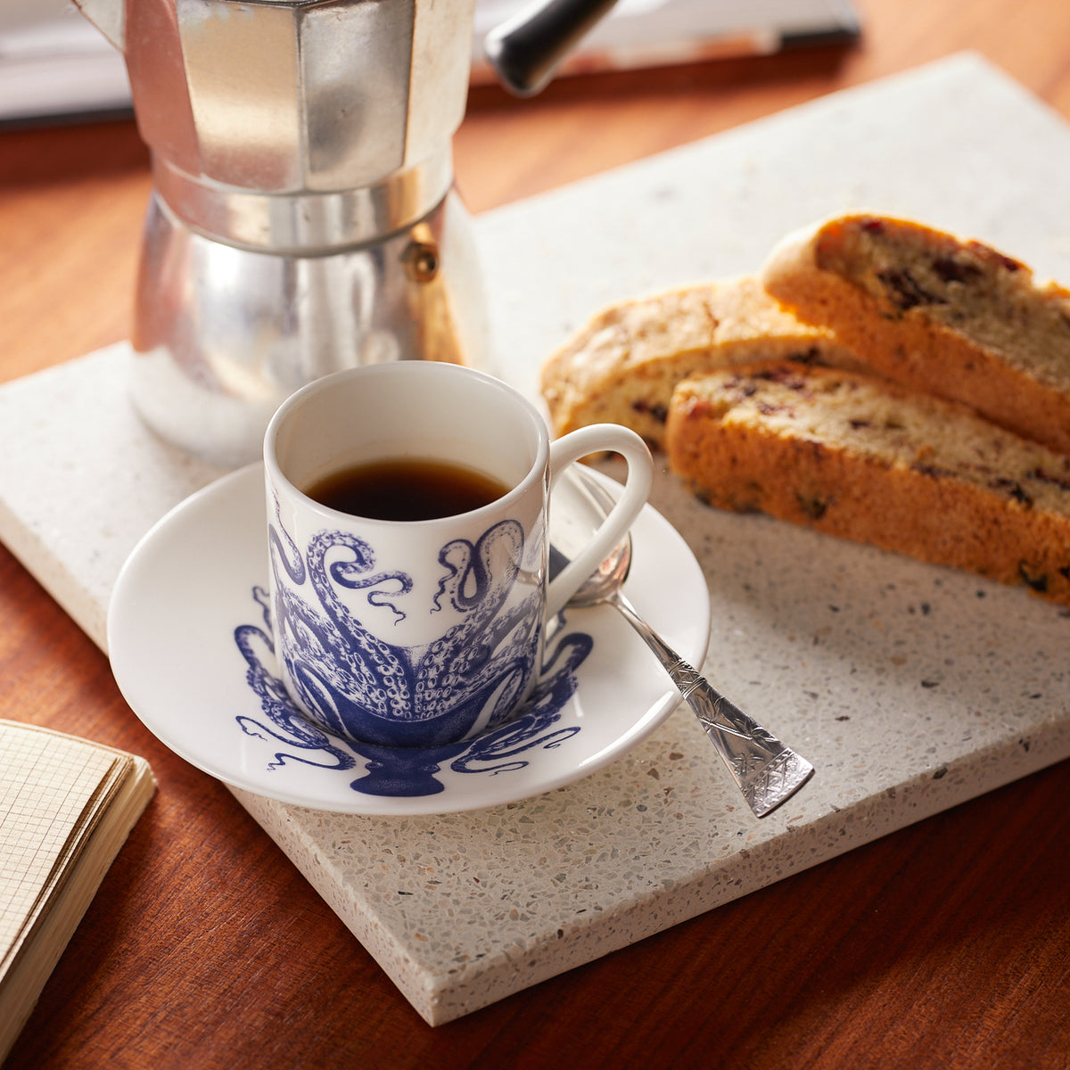 A set from Caskata, the Lucy Espresso Cups &amp; Saucers, Set of 2, adorned with a blue octopus design and accompanied by silver spoons, sits on a countertop. Next to it are two pieces of biscotti and an espresso maker in the background.