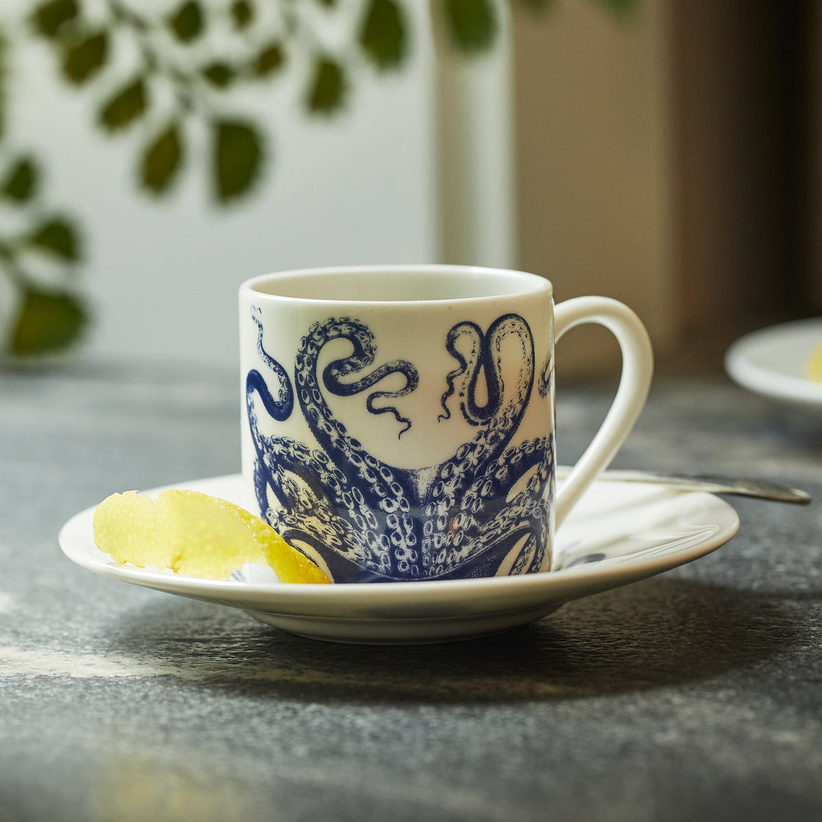 A Caskata Lucy Espresso Cups &amp; Saucers, Set of 2 with a blue octopus design sits on a saucer, holding a lemon slice, with green plant leaves in the background.