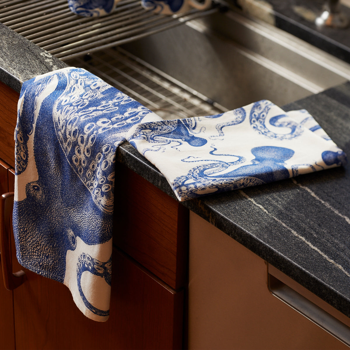 A set of Lucy Kitchen Towels, Set of 2 by Caskata adds a whimsical touch to the kitchen. One cotton tea towel with a blue octopus design is draped over the handle of a wooden cabinet beside the sink, while another rests playfully on the countertop. These delightful kitchen companions bring both style and function to your space.