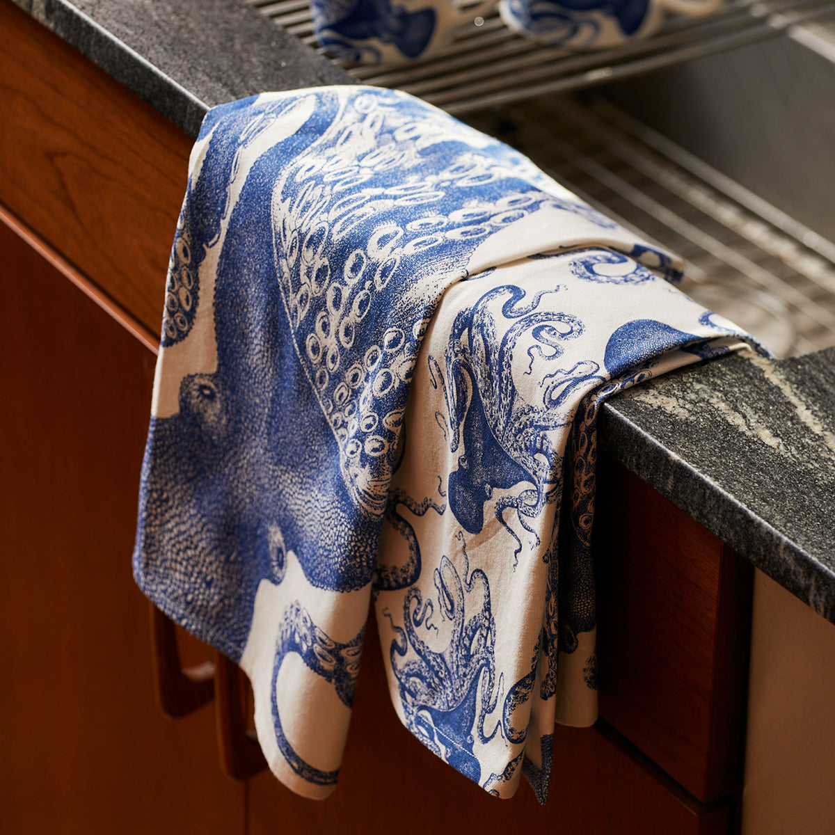 A blue and white **Lucy Kitchen Towel** from the **Caskata** set, one of your favorite kitchen companions, hangs over the edge of a countertop next to a dish drying rack.