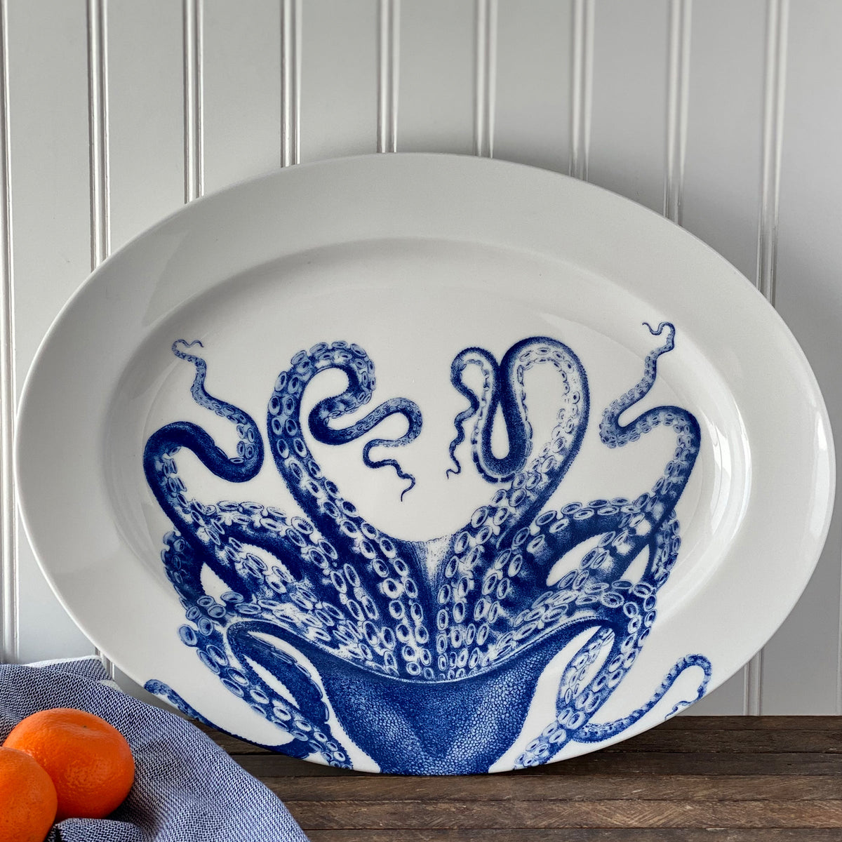 Lucy Oval Rimmed Platter by Caskata Artisanal Home featuring a blue octopus design. The platter is placed on a wooden surface with two small oranges and a blue cloth to the left. Background has white vertical wall panels.
