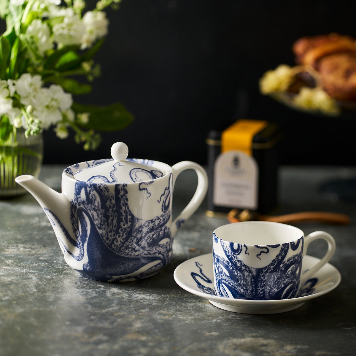 A bone china white teapot and Lucy Cups &amp; Saucers, Set of 2 by Caskata with blue designs sit gracefully on a gray table. In the background, a vase with flowers, a tin of tea, and a plate of pastries complete the charming scene.