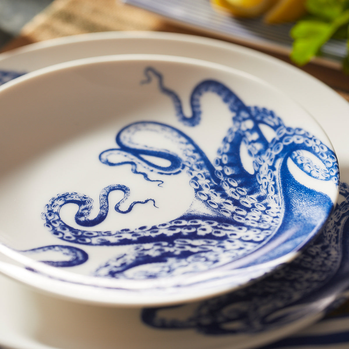 A creamy white premium porcelain bowl with a detailed blue octopus design on its inside surface is stacked on Caskata Artisanal Home&#39;s Lucy Coupe Salad Plates. This contemporary shaped set is both dishwasher and microwave safe.
