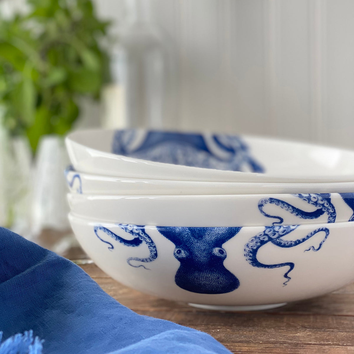 Three Lucy Entrée Bowls by Caskata Artisanal Home with blue designs are stacked on a wooden surface. A blue cloth is partially visible in the foreground, with green leaves and a glass bottle blurred in the background, highlighting the craftsmanship of these premium porcelain pieces.