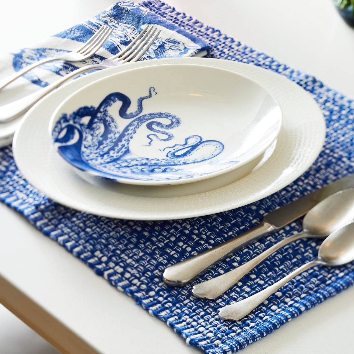 Dining table setting with a blue and white octopus-themed bowl in a contemporary shape, a Lucy Coupe Salad Plate from Caskata Artisanal Home, silverware, and a folded blue patterned napkin, all placed on a blue woven placemat. All items are both dishwasher and microwave safe.