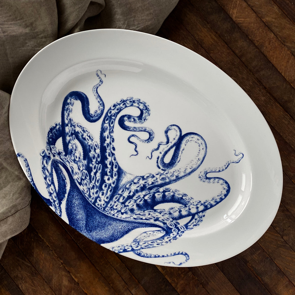 A high-fired porcelain oval dish, the Lucy Oval Rimmed Platter by Caskata Artisanal Home, featuring a blue octopus illustration on the inside, placed on a wooden surface with gray fabric partially visible in the background.