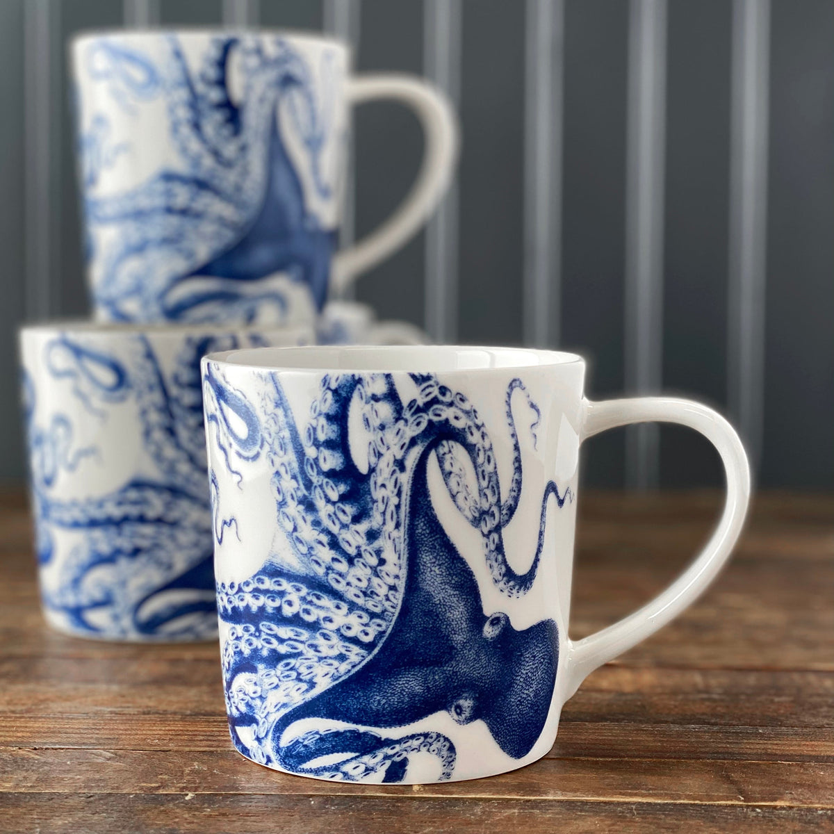 Three white Lucy Mugs by Caskata Artisanal Home with blue illustrations are stacked on a wooden surface, set against a blurred background. These dishwasher and microwave safe mugs add a touch of the ocean to your kitchen.