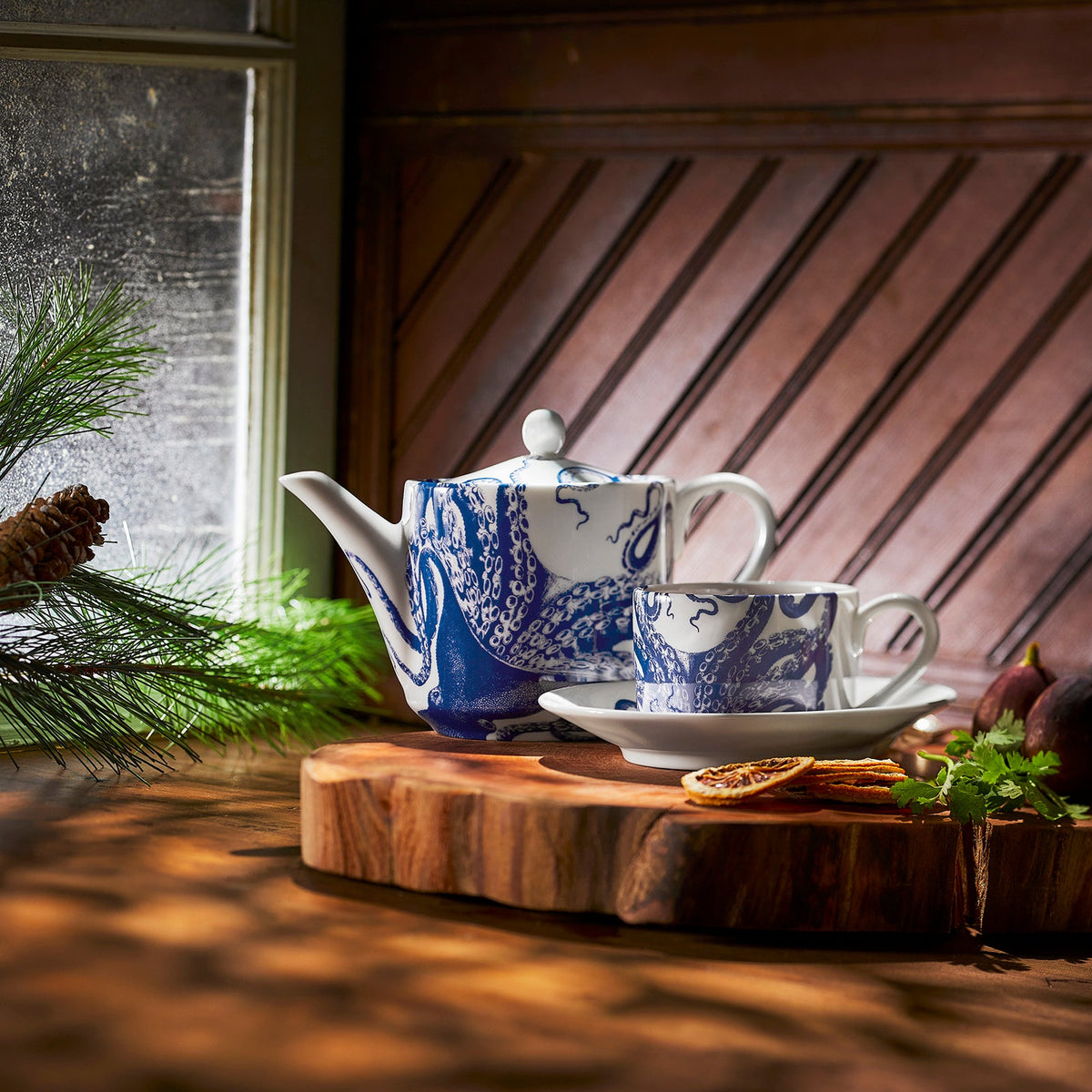Blue and white teapot made of bone china with matching Lucy Cups &amp; Saucers, Set of 2 by Caskata on a wooden tray, placed near a window with sunlight streaming in and greenery in the foreground.