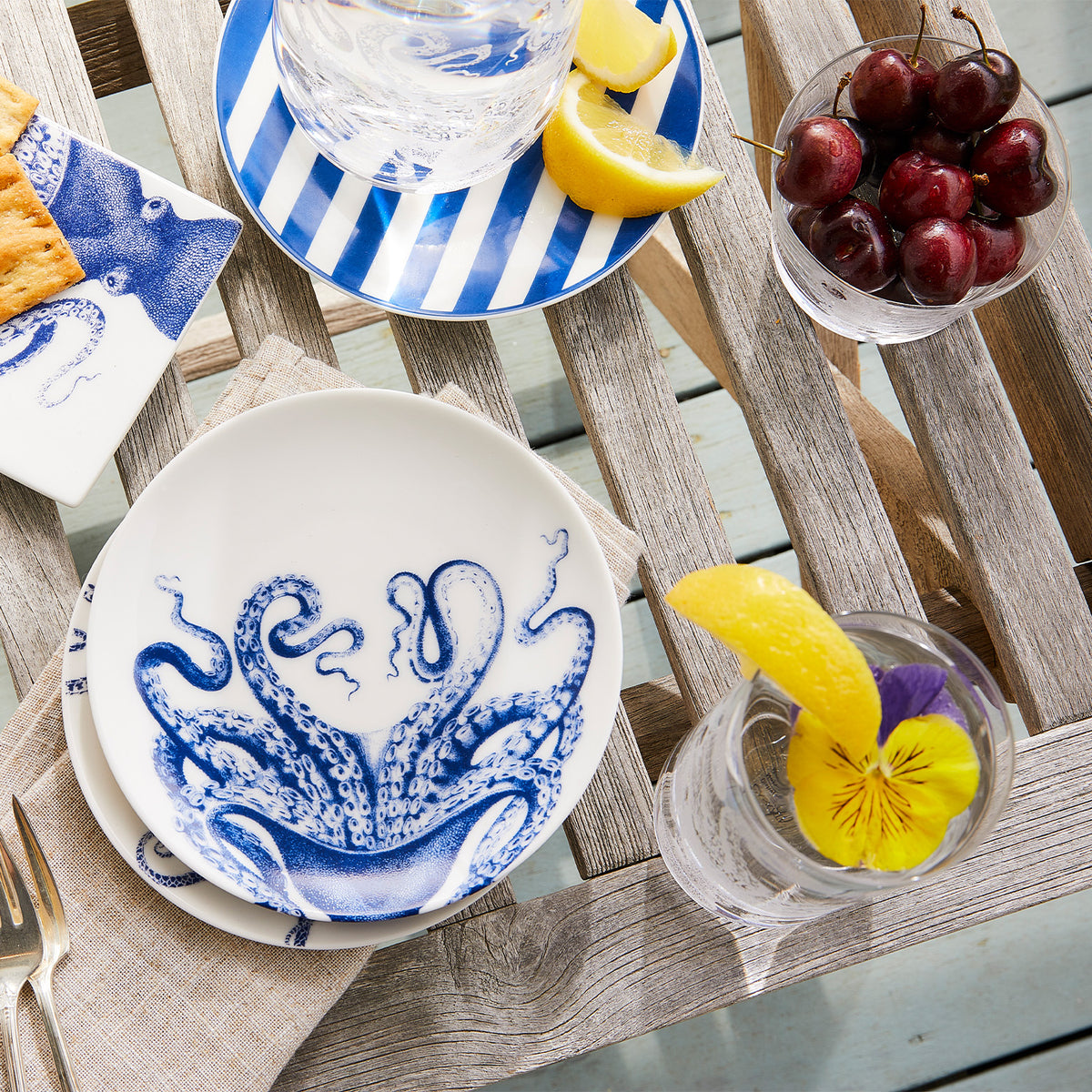 A table with a wooden surface featuring Beach Towel Stripe Small Plates by Caskata Artisanal Home, a bowl of cherries, a glass with a lemon slice and purple flower, and a plate with lemon wedges beside a glass of water.