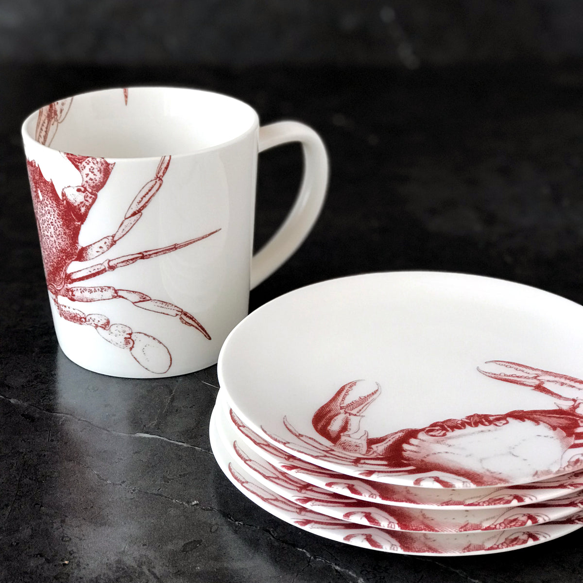 A white mug and a stack of Crab Red Small Plates, each adorned with red crab illustrations, are displayed on a dark surface, showcasing their heirloom-quality dinnerware in lead-free porcelain from Caskata Artisanal Home.