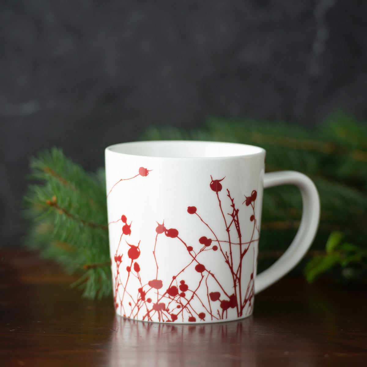 A generously sized, creamy white porcelain Caskata Artisanal Home Winterberries Mug with a red, floral branch design sits on a wooden surface, with green foliage in the background.