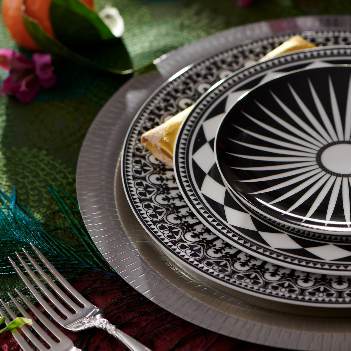 A close-up of a dining table setting featuring ornate black and white geometric pattern plates, specifically the Fez Rimmed Salad Plate by Caskata Artisanal Home, a silver charger, and silverware. The sophisticated global feel is accentuated by a decorative peacock feather and flowers nearby, perfectly complementing the bold geometrics.