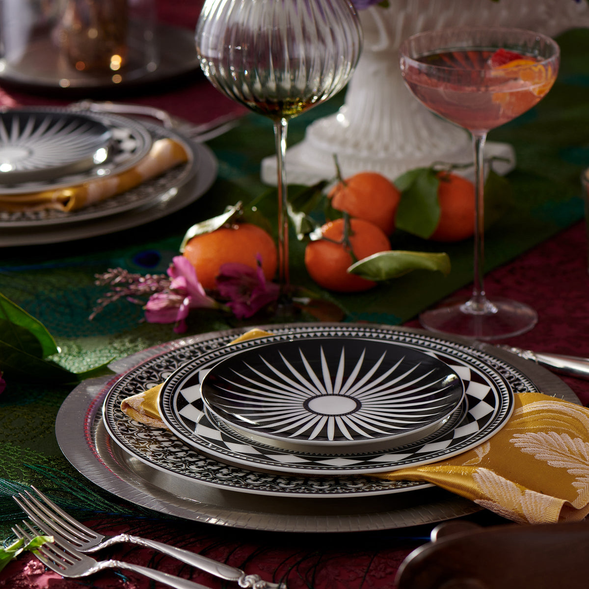 A dinner table setting with Caskata Artisanal Home&#39;s Fez Rimmed Salad Plates, gold utensils, and orange napkins. A cocktail glass and oranges are also on the table, with flowers and greenery adding sophisticated global decor.