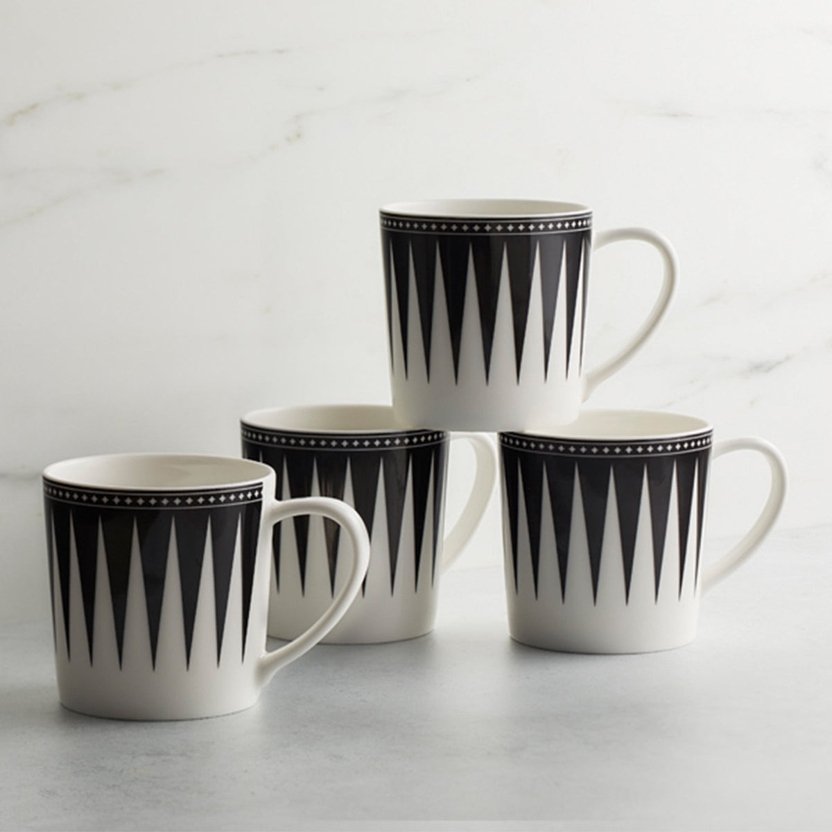 Four high-fired porcelain Marrakech Mugs by Caskata Artisanal Home with black geometric triangle patterns, placed on a light grey surface against a white background. These Art Deco inspired Marrakech Mugs bring an elegant touch to any setting.