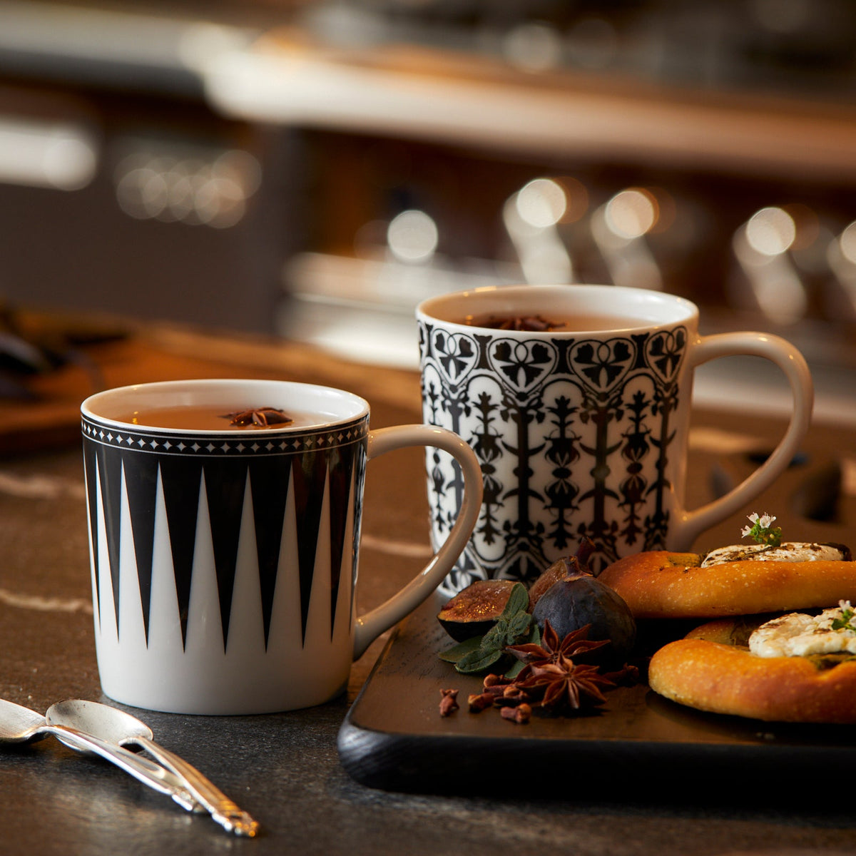 Two Marrakech Mugs from Caskata Artisanal Home with black and white patterns, crafted from high-fired porcelain, are filled with tea and placed on a table next to a tray with pastries, figs, and garnishes. Silver spoons rest nearby. The background shows a blurred kitchen setting.