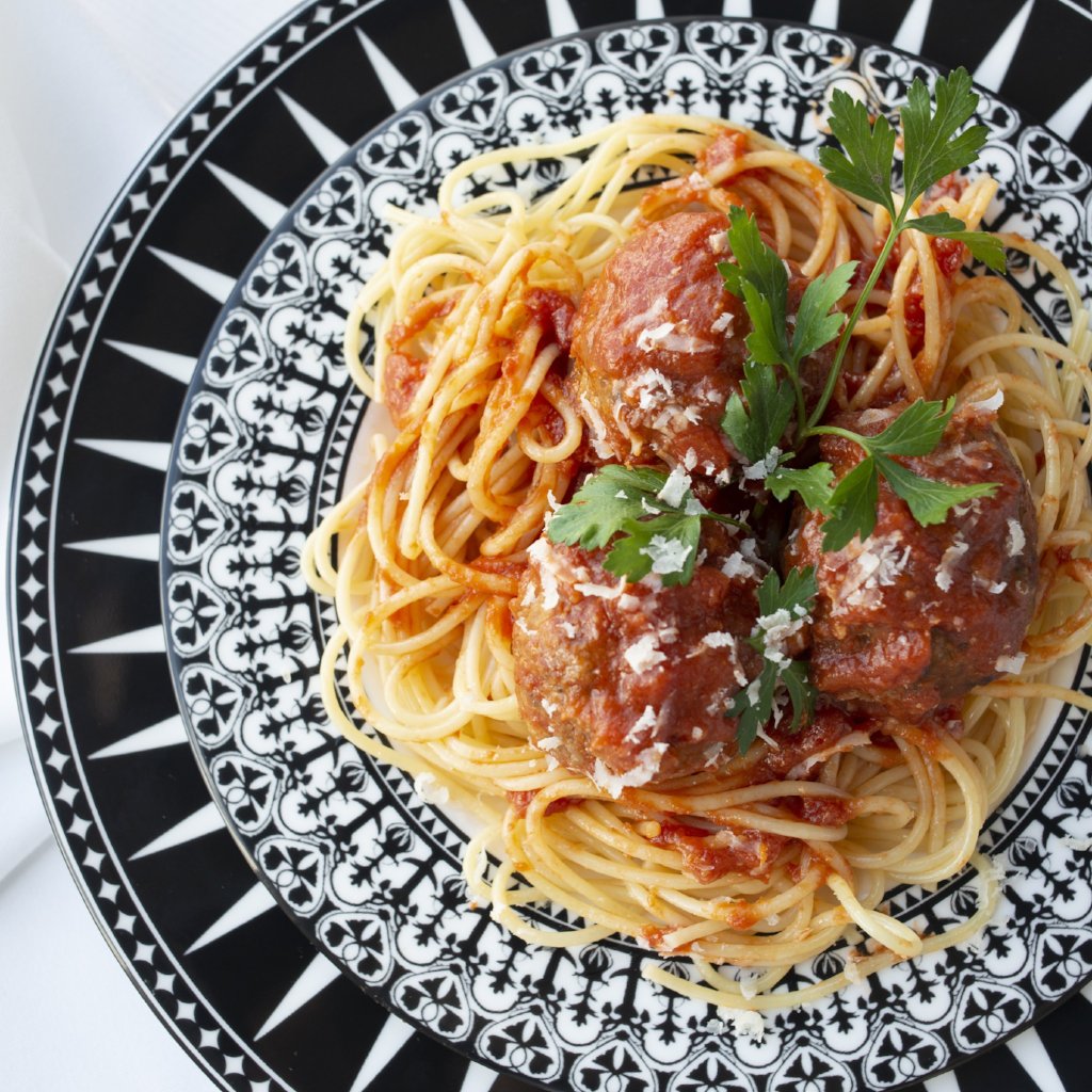 A plate of spaghetti topped with three meatballs in tomato sauce, garnished with grated cheese and fresh parsley, served on a decorative black and white patterned Caskata Artisanal Home Casablanca Rimmed Salad Plate.