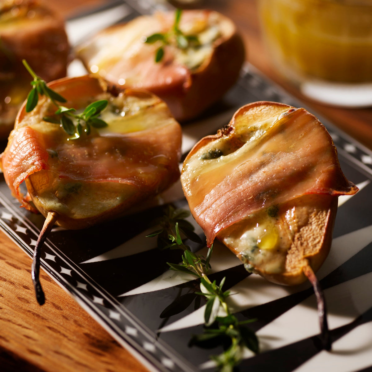 Slices of pears wrapped in bacon with melted cheese and herbs on an elegant Caskata Marrakech Medium Sushi Tray, Set of 2, resting on a wooden surface with a blurred glass jar in the background.