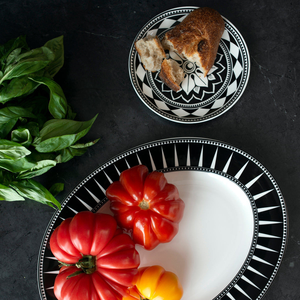 Patterned **Fez Small Plates by Caskata Artisanal Home** featuring bold geometrics on a dark surface, one holding large tomatoes and another with a piece of bread; fresh basil is placed to the side.