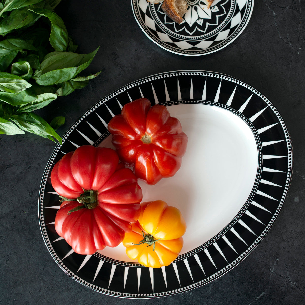 Three heirloom tomatoes, two red and one yellow, rest on a Caskata Artisanal Home Marrakech Oval Rimmed Platter with fresh basil leaves on the side.