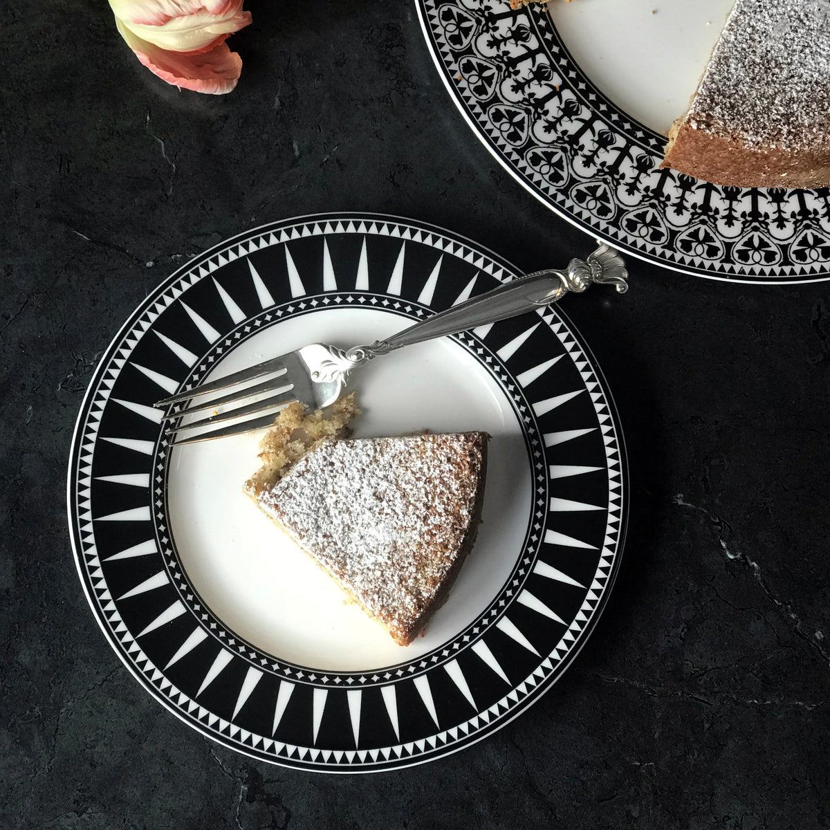 A slice of cake dusted with powdered sugar on a Marrakech Rimmed Salad Plate by Caskata Artisanal Home with a fork, beside a similar plate holding the remaining cake. A flower is partially visible in the corner.