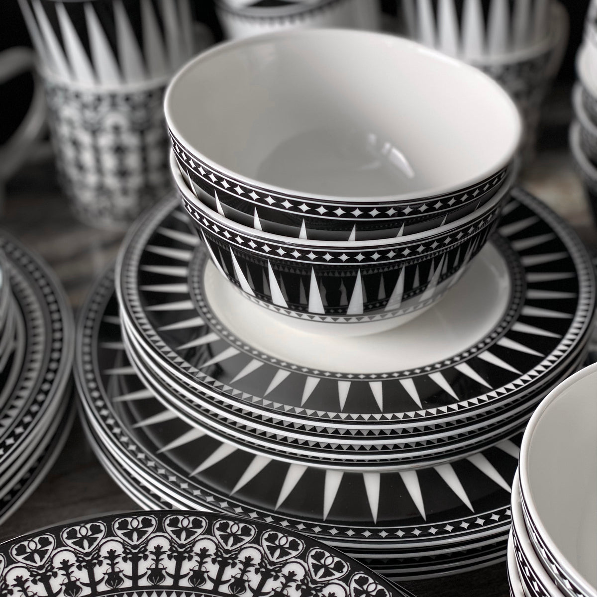 A stack of Art Deco inspired ceramics, featuring black and white patterned dishes including the Marrakech Cereal Bowl from Caskata Artisanal Home and plates with intricate geometric designs, displayed together.