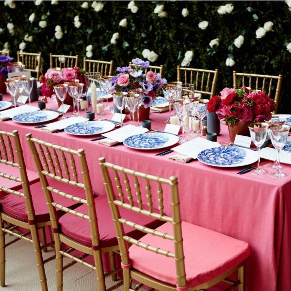 Elegant dining setup with pink tablecloths, premium porcelain Caskata Artisanal Home Peony Coupe dinnerware, gold chairs, and floral centerpieces, under a canopy of white flowers.