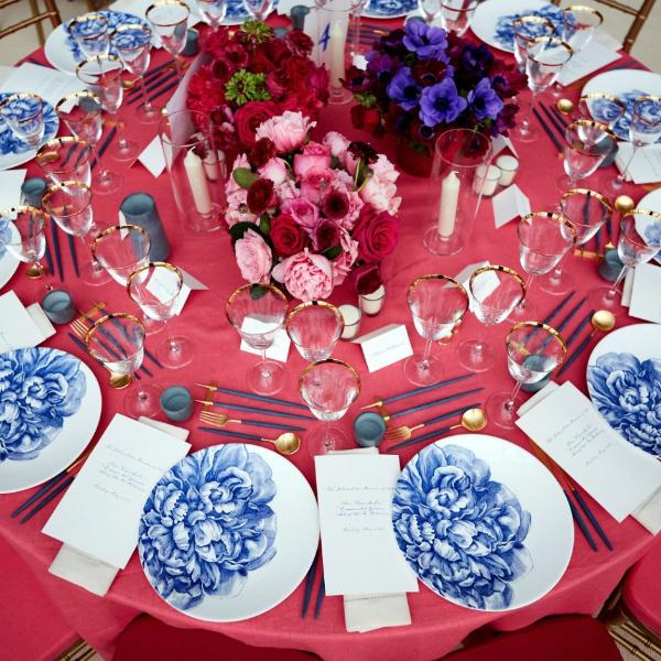 Elegant table setting with red tablecloth, Caskata Artisanal Home&#39;s Peony Coupe Dinner Plate, blue napkins decorated with flower designs, and arranged place settings with name cards.