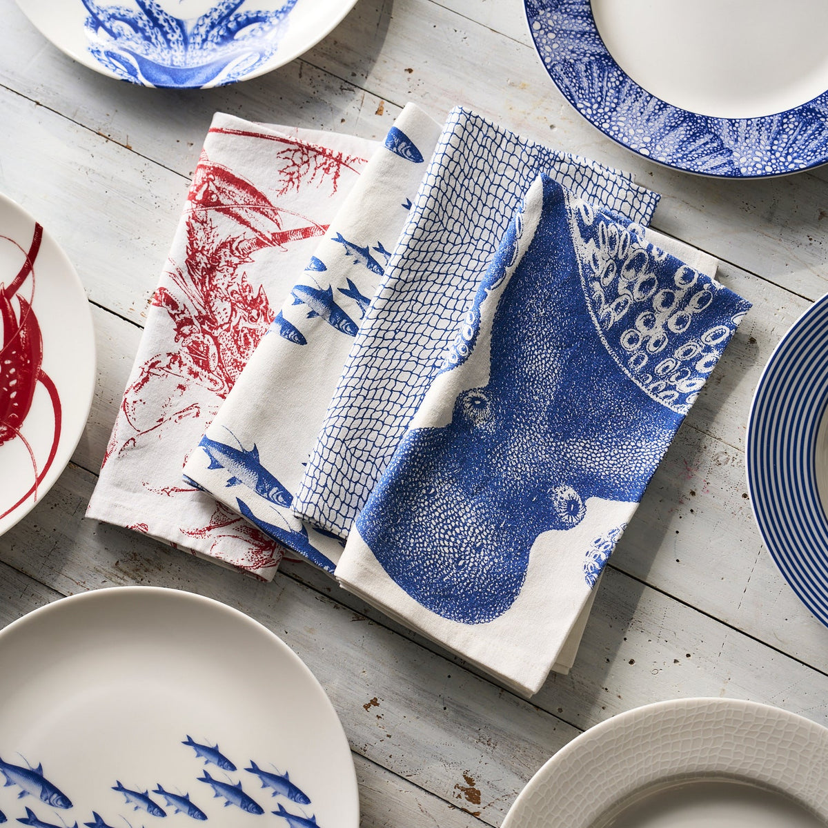 Various blue and white patterned plates and folded tea towels are arranged on a wooden surface. The blue and white Lucy Dinner Napkins, Set of 4 by Caskata feature different marine-themed designs, perfect for any coastal-inspired setting.